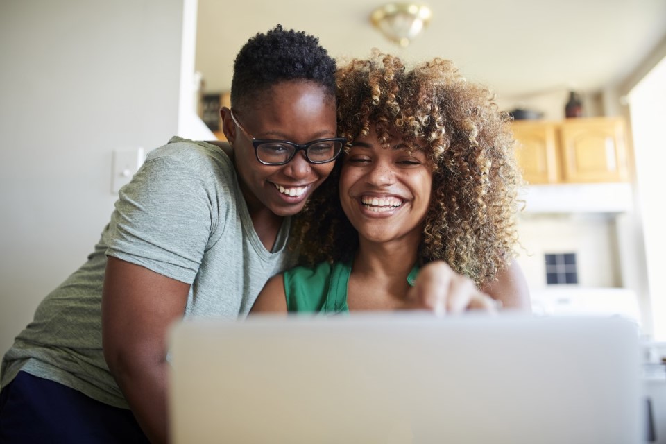 Laughing Black women hugging and using laptop.