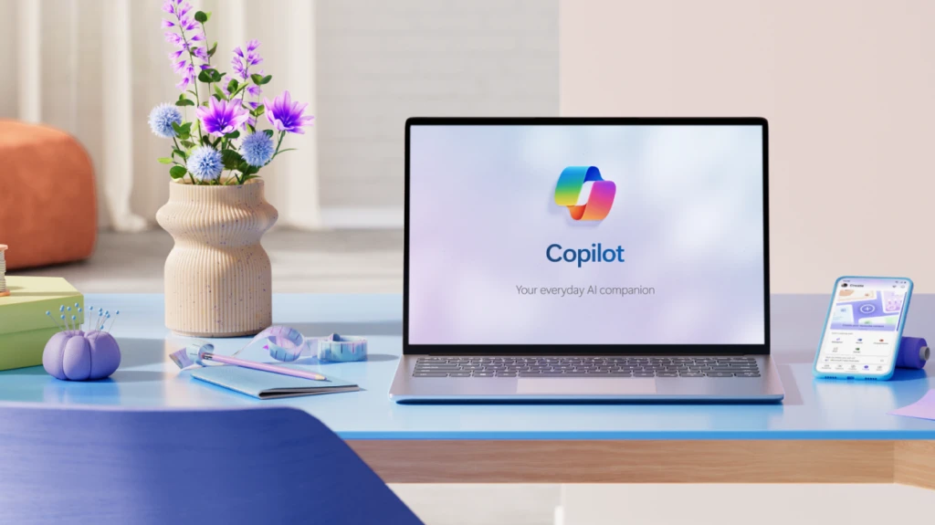 A laptop with the Copilot icon on screen on a blue desk with a vase of purple flowers, sewing cushion, and cell phone.