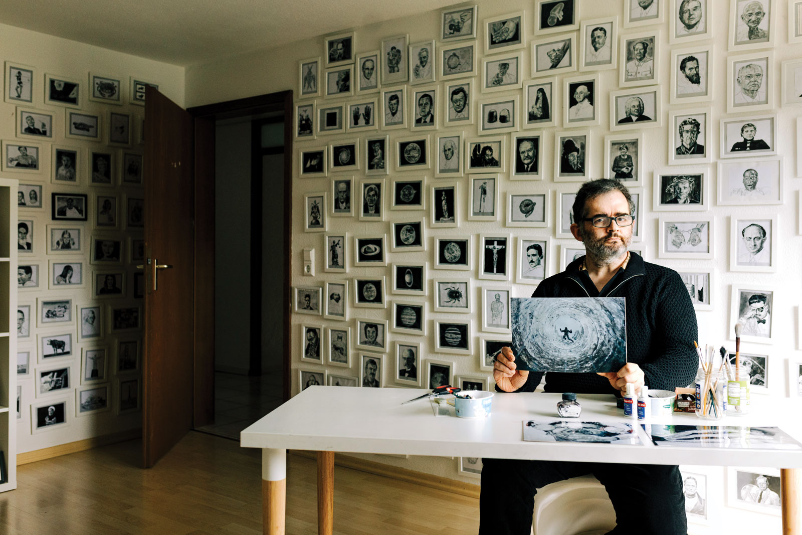 Thomas Müller sits at a desk and holds up a painting. The painting is in black and white and depicts a silhouetted figure falling into a deep hole. Müller's expression is serious. The walls behind him are filled from floor to ceiling with more black and white artwork in white frames.