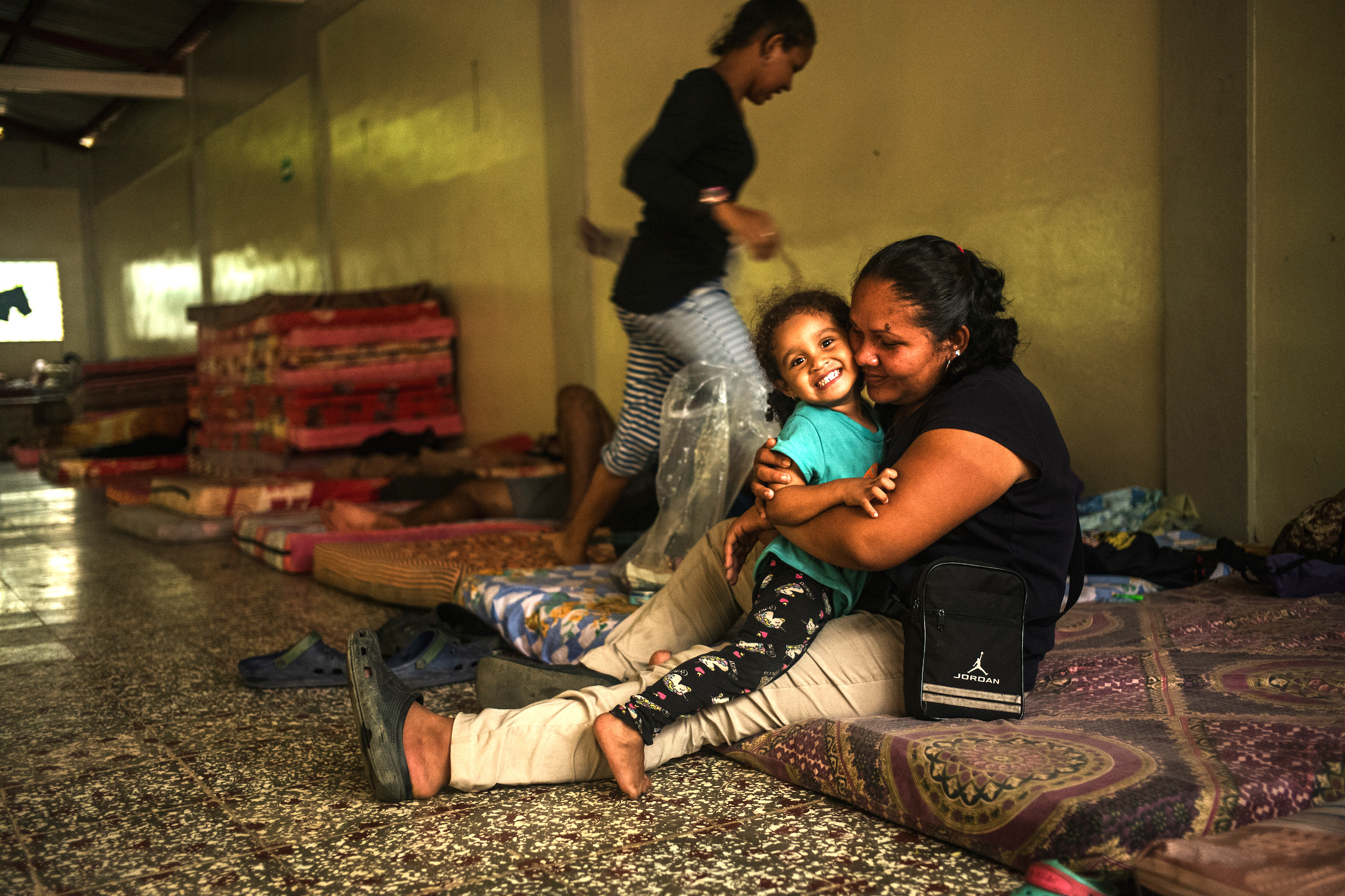A woman and a smiling girl sit on a mattress on the ground.